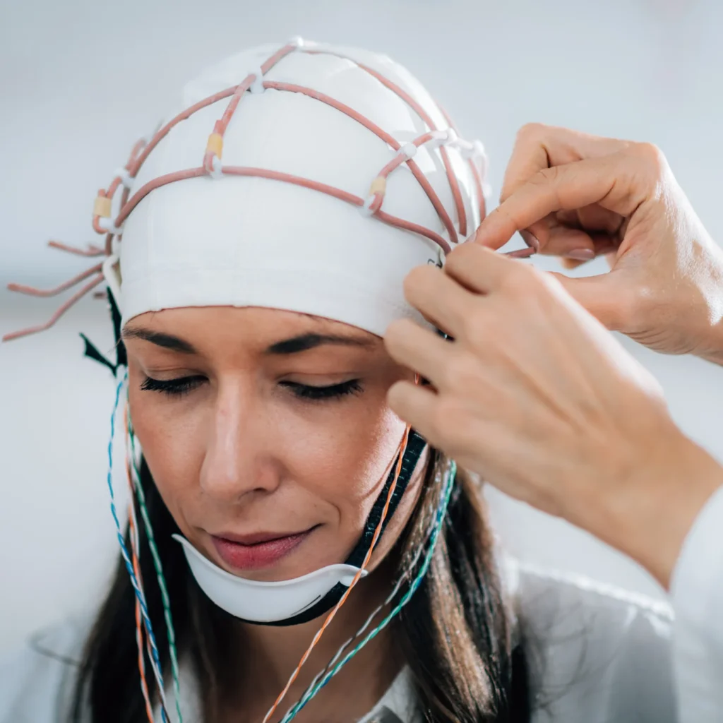 Person wearing an EEG cap with electrodes attached, while another person adjusts it.