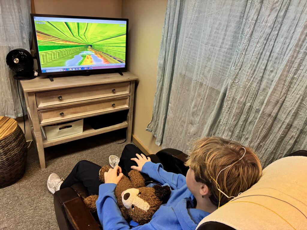 A person with electrodes on their head watches a screen displaying a virtual tunnel while holding a stuffed animal, seated in a room with light curtains and a wooden desk nearby.